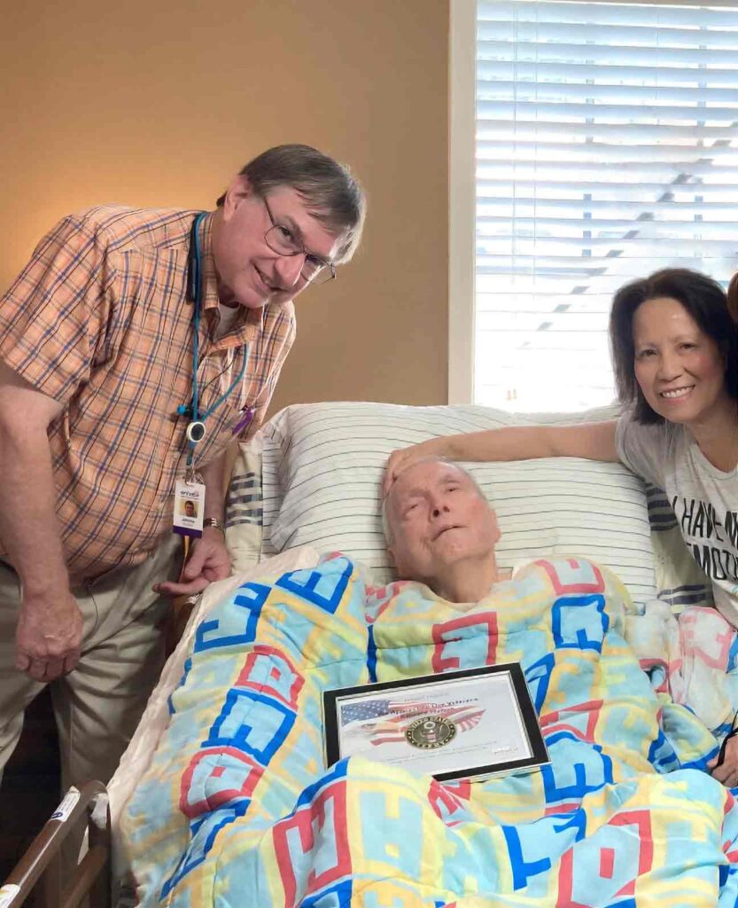 Veteran hospice patient laying in bed with his loved ones and clinician on either side of him.