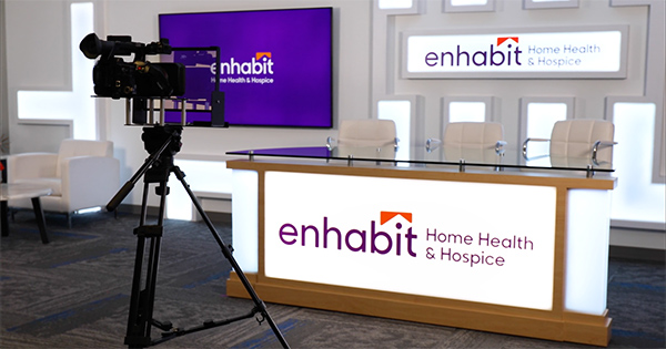 This is a picture of the studio at the Enhabit Enrichment Community (EEC). It shows 3 white chairs behind a white table with the Enhabit logo on it. A professional camera set up is in the front of the frame as well.