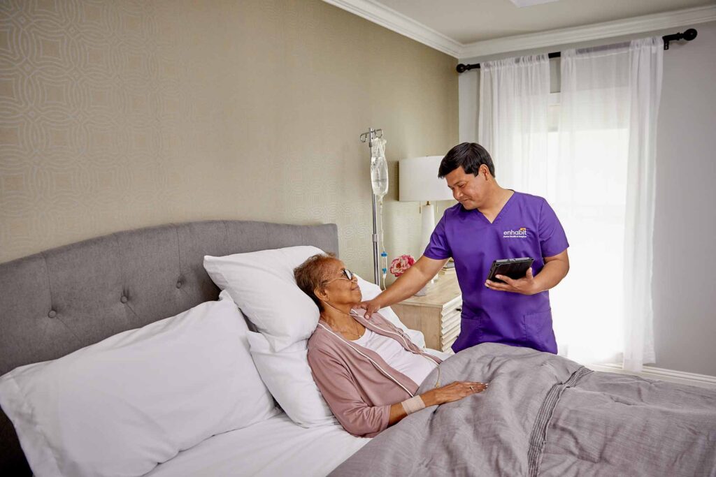 The hospice provider, an Enhabit clinician, puts his hand on a hospice patients shoulder while she lies in bed, hooked up to an IV.